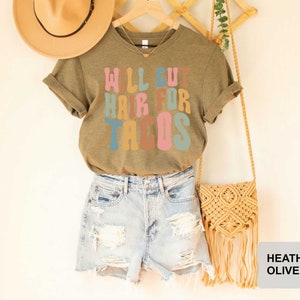 May include: A heather olive t-shirt with the text "Will cut hair for tacos" in colorful letters. The shirt is modeled on a person wearing denim shorts and a straw hat.