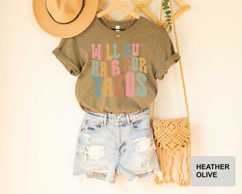 May include: A heather olive t-shirt with the text "Will cut hair for tacos" in colorful letters. The shirt is modeled on a person wearing denim shorts and a straw hat.
