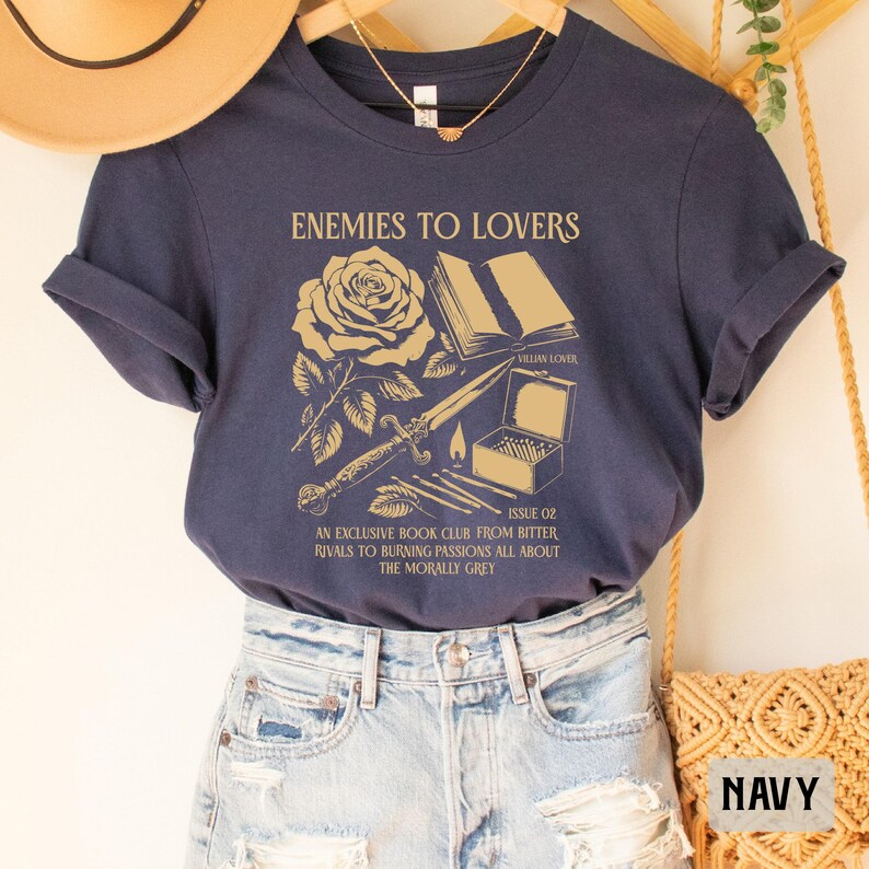 May include: A navy blue t-shirt with a gold graphic design featuring a rose, a book, a dagger, and matches. The text on the shirt reads "Enemies to Lovers", "Villain Lover", "Issue 02", "An Exclusive Book Club From Bitter Rivals to Burning Passions All About The Morally Grey".