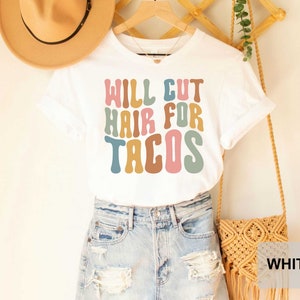 May include: White t-shirt with the text "WILL CUT HAIR FOR TACOS" in colorful, retro-style lettering.