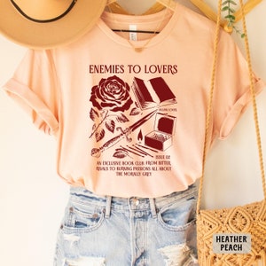 May include: Peach colored t-shirt with a red graphic design featuring a rose, a book, a dagger, matches, and the text "Enemies to Lovers" and "Issue 02 An Exclusive Book Club From Bitter Rivals to Burning Passions All About The Morally Grey".