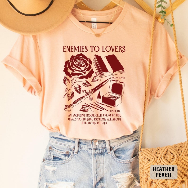 May include: Peach colored t-shirt with a red graphic design featuring a rose, a book, a dagger, matches, and the text "Enemies to Lovers" and "Issue 02 An Exclusive Book Club From Bitter Rivals to Burning Passions All About The Morally Grey".