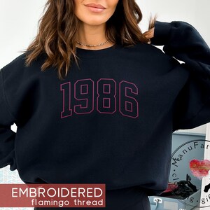 May include: A black long-sleeve crewneck sweatshirt featuring the embroidered text 1986 in a dark red, varsity-style font across the chest. The garment has a classic fit with ribbed cuffs and a ribbed neckline, showcasing a clean, simple design.