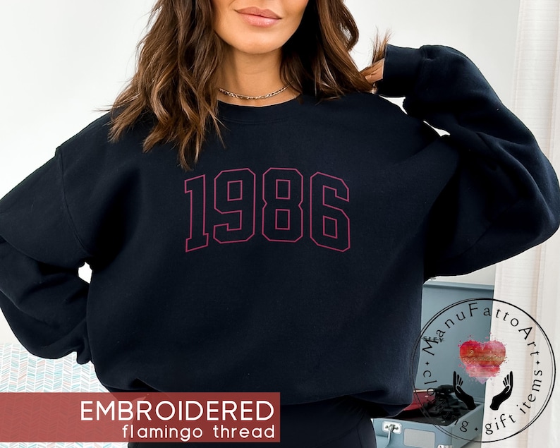 May include: A black long-sleeve crewneck sweatshirt featuring the embroidered text 1986 in a dark red, varsity-style font across the chest. The garment has a classic fit with ribbed cuffs and a ribbed neckline, showcasing a clean, simple design.