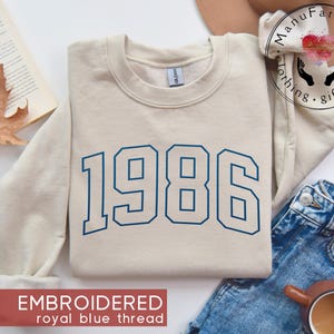 May include: A beige crewneck sweatshirt featuring the year 1986 embroidered in royal blue thread across the chest. The garment has long sleeves, a ribbed collar, and a Gildan brand label visible at the inner neckline.