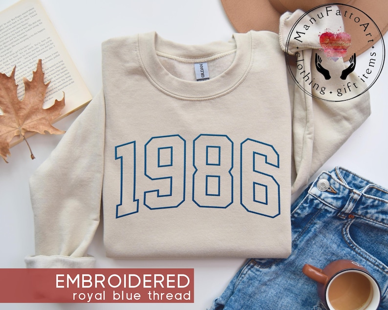 May include: A beige crewneck sweatshirt featuring the year 1986 embroidered in royal blue thread across the chest. The garment has long sleeves, a ribbed collar, and a Gildan brand label visible at the inner neckline.