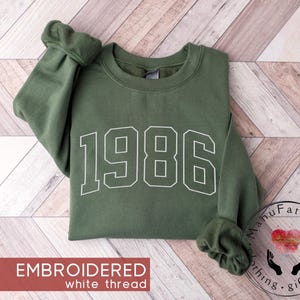 May include: An olive green crewneck sweatshirt featuring the year 1986 embroidered in white thread across the chest. The garment has long sleeves with ribbed cuffs and a ribbed neckline, showcasing a clean, athletic-style font for the numbers.