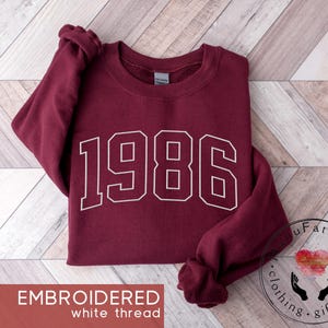 May include: A maroon long-sleeve crewneck sweatshirt featuring the number 1986 embroidered in white thread across the chest. The garment has a ribbed collar, cuffs, and hem, with a Gildan brand label visible inside the neckline of the shirt.