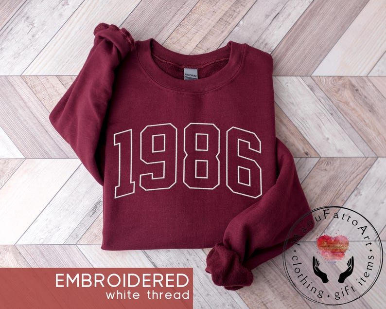 May include: A maroon long-sleeve crewneck sweatshirt featuring the number 1986 embroidered in white thread across the chest. The garment has a ribbed collar, cuffs, and hem, with a Gildan brand label visible inside the neckline of the shirt.