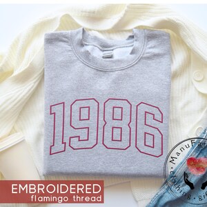 May include: A light gray crewneck sweatshirt featuring the year 1986 embroidered in a bold, dark red varsity-style font across the chest. The garment has a classic ribbed neckline and long sleeves, showcasing a clean and simple design aesthetic.