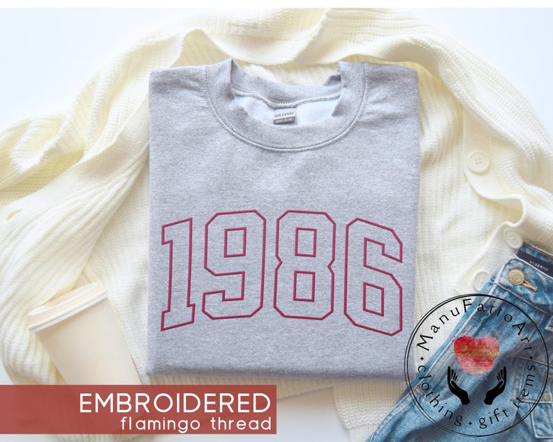 May include: A light gray crewneck sweatshirt featuring the year 1986 embroidered in a bold, dark red varsity-style font across the chest. The garment has a classic ribbed neckline and long sleeves, showcasing a clean and simple design aesthetic.