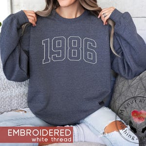 May include: A dark charcoal gray crewneck sweatshirt featuring the year 1986 embroidered in a white collegiate-style font across the chest. The garment has long sleeves, a ribbed neckline, and a soft, heathered fabric texture throughout.