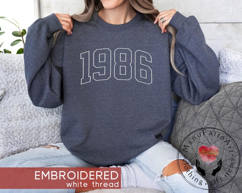 May include: A dark charcoal gray crewneck sweatshirt featuring the year 1986 embroidered in a white collegiate-style font across the chest. The garment has long sleeves, a ribbed neckline, and a soft, heathered fabric texture throughout.