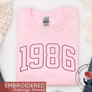 May include: A light pink crewneck sweatshirt featuring the year 1986 embroidered in a dark maroon varsity-style font across the chest. The garment has a Gildan brand label at the inner neckline and is made of a soft, solid-colored fabric.