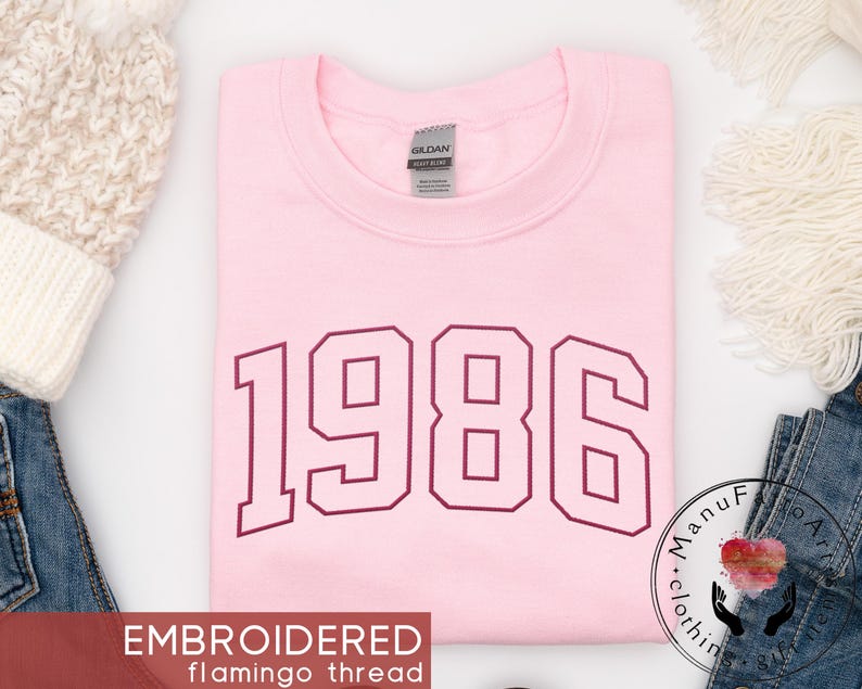 May include: A light pink crewneck sweatshirt featuring the year 1986 embroidered in a dark maroon varsity-style font across the chest. The garment has a Gildan brand label at the inner neckline and is made of a soft, solid-colored fabric.