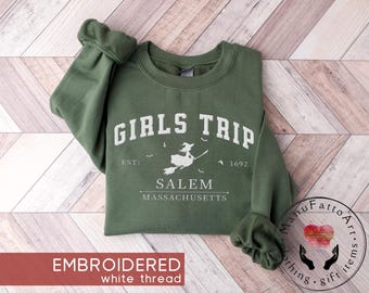 May include: An olive green crewneck sweatshirt featuring white embroidered text that reads GIRLS TRIP EST: 1692 SALEM MASSACHUSETTS. The design includes a central graphic of a witch flying on a broomstick surrounded by small bats.