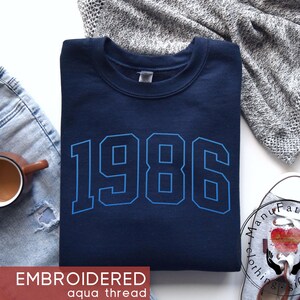 May include: A navy blue crewneck sweatshirt featuring the year 1986 embroidered in a bold, light blue athletic font across the chest. The garment is displayed with a pair of light wash distressed jeans, a coffee mug, and a gray knit fabric.