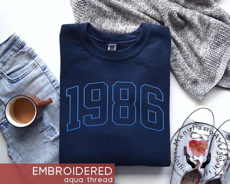 May include: A navy blue crewneck sweatshirt featuring the year 1986 embroidered in a bold, light blue athletic font across the chest. The garment is displayed with a pair of light wash distressed jeans, a coffee mug, and a gray knit fabric.