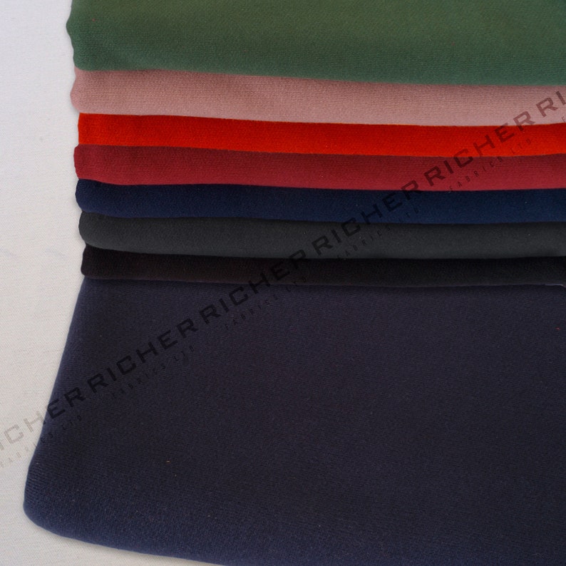 Op de afbeelding: Een stapel stofstalen in verschillende kleuren, waaronder groen, roze, rood, bordeauxrood, donkerblauw en zwart. De stof lijkt een zacht, getextureerd materiaal te zijn.