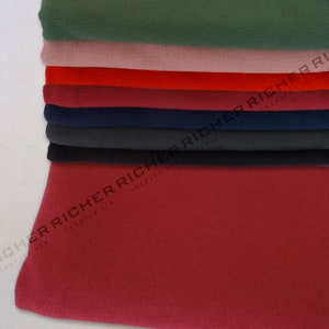 Op de afbeelding: Een stapel stofstalen in verschillende kleuren, waaronder groen, roze, rood, bordeauxrood, donkerblauw, zwart en donkerrood. De stof is glad en heeft een subtiele textuur. De stalen zijn netjes gevouwen en op elkaar gestapeld. De tekst "RICHERRICHER FABRICS" is zichtbaar op het onderste staal.