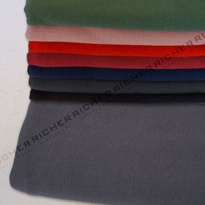 Op de afbeelding: Een stapel stofstalen in verschillende kleuren, waaronder groen, roze, rood, bordeauxrood, donkerblauw en grijs. De stof is netjes gevouwen en het onderste staal is het grootste.
