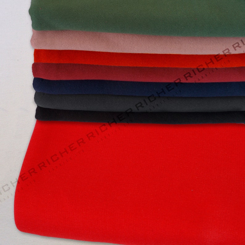 Op de afbeelding: Een stapel stofstalen in verschillende kleuren, waaronder rood, groen, zwart, donkerblauw en bordeauxrood. De stalen zijn netjes gevouwen en in een verticale stapel gerangschikt.