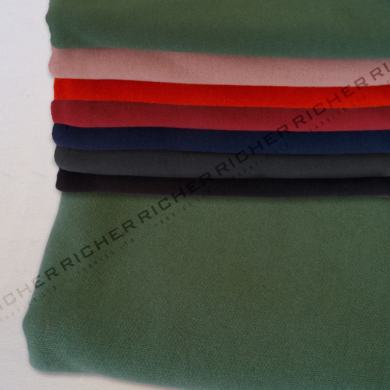 Op de afbeelding: Een stapel stofstalen in verschillende kleuren, waaronder groen, roze, rood, bordeauxrood, donkerblauw en zwart. De stof is glad en heeft een lichte glans.