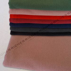 Op de afbeelding: Een stapel stofstalen in verschillende kleuren, waaronder groen, roze, rood, bordeauxrood, donkerblauw, grijs en zwart. De stof is glad en heeft een subtiele textuur.