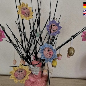 Może przedstawiać: Wazon wypełniony gałęziami ozdobionymi dzianinowymi kwiatami i pisankami wielkanocnymi. Kwiaty są w odcieniach różowego, żółtego i niebieskiego, a pisanki są złote i białe.