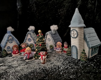 May include: A crocheted Christmas village scene with houses, a church, a Christmas tree, and small figures. The houses are white with blue roofs and have white smoke coming from the chimneys. The church has a clock tower with Roman numerals. The Christmas tree is green with gold ornaments and a red star on top. The figures are wearing red, blue, and purple clothing. The scene is set on a black background with white snow.