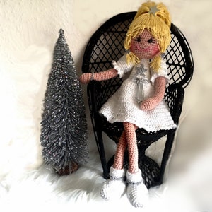 Könnte beinhalten: Eine gehäkelte Puppe mit blonden Haaren und einem weißen Kleid sitzt auf einem schwarzen Korbstuhl. Die Puppe trägt weiße Schuhe und hat ein Schneeflockenmuster auf ihrem Kleid. Im Hintergrund steht ein silberner Glitzer-Weihnachtsbaum.