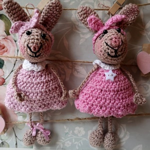 Könnte beinhalten: Zwei gehäkelte rosa Hasen mit weißen Akzenten, die an einer Schnur mit Wäscheklammern hängen. Die Hasen haben braune Gesichter und rosa Kleider.