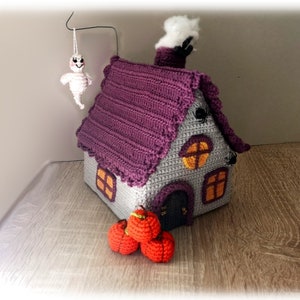 May include: A crocheted Halloween house with a purple roof, gray walls, and orange windows. There are three small orange pumpkins in front of the house and a white ghost hanging from the ceiling.