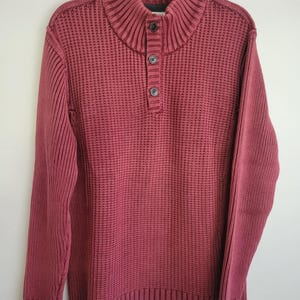 以下が含まれることがあります： リブ編みのテクスチャとボタン留めのハイネックが特徴の、マルーン色のセーター。長袖セーターは、白い背景に黒いハンガーに掛けられています。セーターには前面に3つのボタンが付いています。