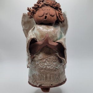May include: A handmade ceramic angel figurine with a star. The angel has a brown face and hair, white wings, and a white and brown robe. The figurine is suspended by a brown string, and is a decorative item.