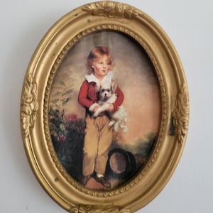 Puede incluir: Retrato ovalado enmarcado de un niño sosteniendo un perro blanco pequeño. El niño lleva una chaqueta roja, pantalones color canela y zapatos marrones. El marco dorado tiene detalles ornamentales.