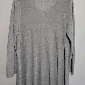 May include: A long-sleeved, gray tunic-style top with a scoop neckline. The fabric has a subtle textured appearance. The hem and cuffs are ribbed. The brand name "Eileen Fisher" is visible at the neckline.