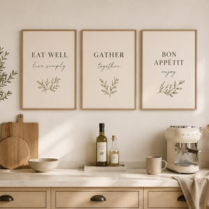 Può includere: Tre stampe incorniciate con testo: "EAT WELL live simply", "GATHER together" e "BON APPÉTIT enjoy". Le stampe sono sopra un piano cucina con bottiglie di olio d'oliva, una macchina per il caffè e utensili di legno.