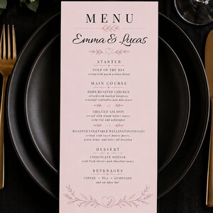 Può includere: Un biglietto del menu di nozze con i nomi "Emma & Lucas". Il menu, in una tonalità rosa tenue, dettaglia un antipasto, piatti principali come pollo arrosto alle erbe e salmone grigliato, dessert e bevande. Decorazioni floreali adornano la carta.