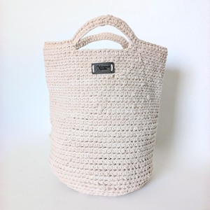May include: A cream-colored, crocheted tote bag with two handles. The bag has a cylindrical shape and a small, rectangular label near the top. The crochet pattern is visible throughout the bag's construction.