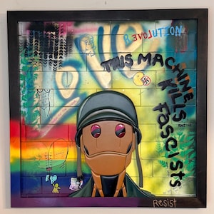 Peut inclure: Une œuvre d'art murale encadrée en techniques mixtes représentant un personnage central en relief portant un casque vert. Le fond est une peinture murale en briques avec des graffitis, incluant le texte "THIS MACHINE KILLS FASCISTS", "LOVE", "REVOLUTION" et "Resist".