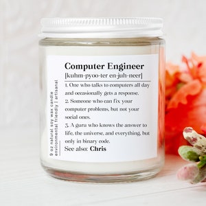 May include: A clear glass jar candle with a white lid and a white label featuring the text Computer Engineer, a phonetic pronunciation, and three humorous definitions. The label also includes the text 9 oz natural soy wax candle.