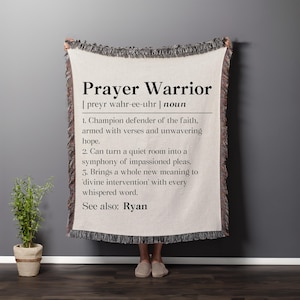 May include: A cream-colored woven throw blanket featuring black text that defines the term Prayer Warrior. The text includes a phonetic pronunciation, a noun label, three numbered definitions, and a see also reference to the name Ryan.