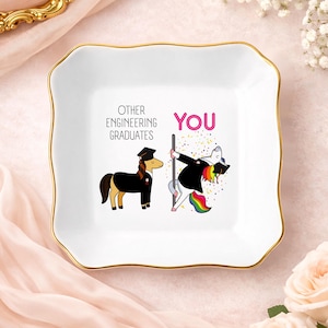 May include: White ceramic trinket dish with a gold rim. The dish features a humorous illustration of a unicorn and a pony, with the text "OTHER ENGINEERING GRADUATES" and "YOU". The unicorn is pole dancing, and the pony is standing still.