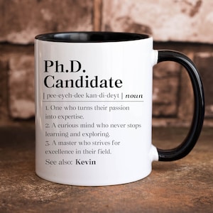 May include: White ceramic mug with a black rim and handle. The mug features a definition of "Ph.D. Candidate" with the phonetic spelling and a list of characteristics. The text at the bottom reads "See also: Kevin."