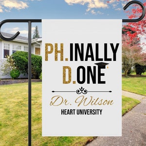 May include: White graduation yard sign with gold glitter text that reads "PH.INALLY D.ONE" with a black graduation cap above the "D". Below the text is "Dr. Wilson HEART UNIVERSITY" in black cursive text.
