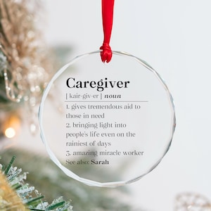 May include: A clear, round ornament with the word "Caregiver" and its pronunciation. The ornament has a red ribbon for hanging and includes a definition with three points. The background is blurred with festive decorations.