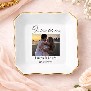 May include: A square white ceramic trinket dish with a gold-trimmed scalloped edge. The center features a printed photograph of a couple on a beach, the text "Our forever starts here.", the names "Lukas & Laura", and the date "05.29.2026".