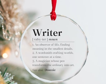 May include: A clear glass ornament with a definition of the word "Writer" printed on it. The definition includes the pronunciation, part of speech, and several definitions. The ornament is hanging from a red ribbon.