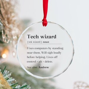 May include: A clear, round ornament with the text "Tech wizard" and a definition. The ornament is suspended by a red ribbon. The definition humorously describes a tech wizard's computer-fixing habits.